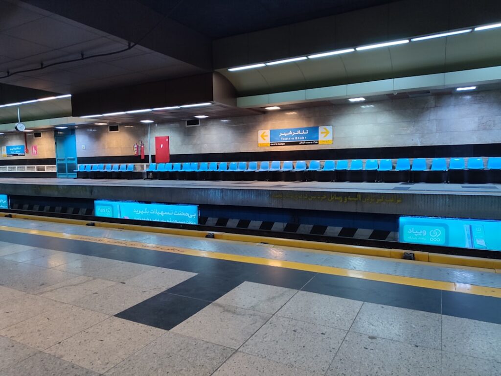Empty subway platform with rows of blue seats and illuminated advertising panels.